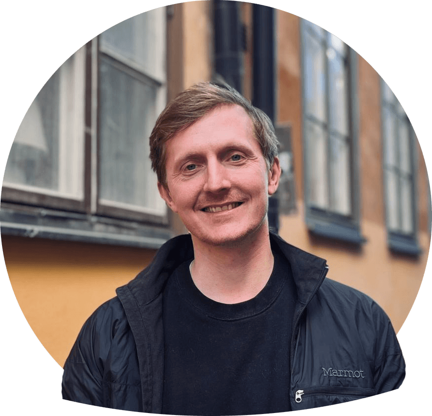 Santino ler mot kameraet foran en gul vegg i Gamla stan, iklädd svart jakke og t-skjorte.